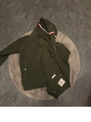 Olivgrön vindjacka från Moncler - Snygg olivgrön vindjacka från Moncler med huva som har coola ränder i rött, vitt och blått. Jackan har dragkedja, tryckknappar och Moncler-logga på ärmen. Perfekt för dig som vill ha en stilren jacka med street-vibe och exklusiv känsla.
