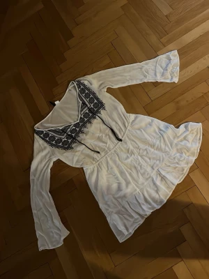 Vit kortklänning med broderi - Supersöt vit kortklänning med mörkblå broderi och snörning vid halsen. Klänningen har långa, lätt utsvängda ärmar och volang nedtill. Perfekt för sommarens alla tillfällen och ger en bohemisk vibe.