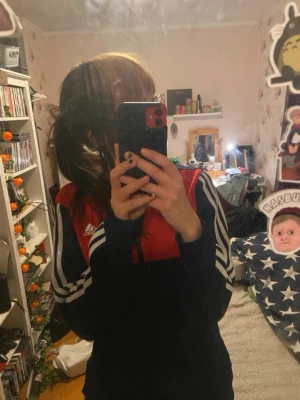 Adidas ziptröja röd och marinblå - Säljer en snygg Adidas ziptröja i marinblått och rött med vita ränder längs ärmarna. Tröjan har hel dragkedja, ståkrage och klassisk Adidas-logga på bröstet. Perfekt till träning eller chill. Materialet är mjukt och skönt, troligtvis polyester.