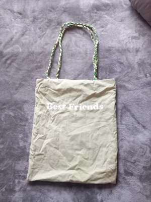Ljusgrön handgjord shoppingväska med flätade handtag - Säljer en handgjord ljusgröntygkasse med texten 'Best Friends' i vitt. Väskan har långa, flätade handtag i grönt, vitt och beige samt gröna detaljer på baksidan. Perfekt för att bära böcker eller småsaker. Enkel och stilren design.