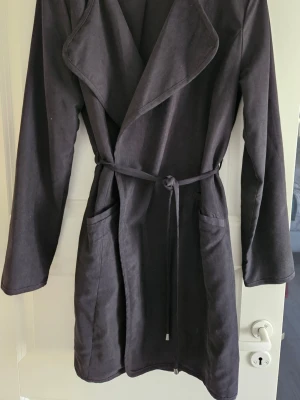 Svart trenchcoat från VILA, storlek M - Säljer en svart trenchcoat från VILA i storlek M. Jackan har ett snyggt omlott-utseende med bälte i midjan och stora fickor framtill. Materialet känns mjukt och lätt, perfekt för lager på lager. Långärmad och klassisk design som funkar till många stilar.