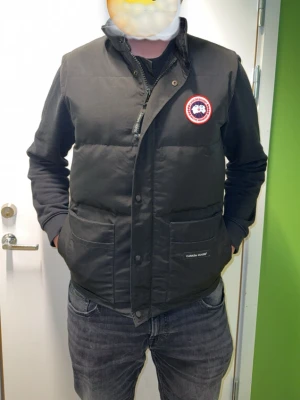 Svart dunväst från Canada Goose - Säljer en svart dunväst från Canada Goose med klassisk logga på bröstet. Västen har dragkedja och tryckknappar framtill, två stora fickor och en hög krage med mjukt foder. Perfekt för lager-på-lager och riktigt snygg till jeans.