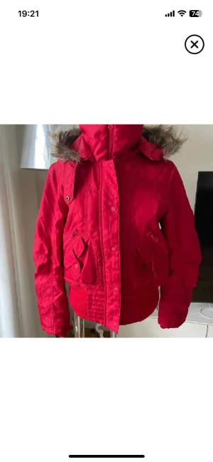 Röd parkajacka med fuskpäls - Säljer en röd parkajacka med huva och fuskpälsdetalj. Jackan har dragkedja och knappar framtill, samt praktiska fickor med både knapp och dragkedja. Ribbad mudd nedtill och vid ärmslut för extra komfort. Perfekt för kyliga dagar.