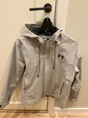 Under Armour jacka - Säljer en ljusgrå vindjacka från Under Armour i storlek M (passar S). Jackan har, svarta detaljer och dragkedja framtill. Perfekt för våran som kommer! Jackan är lite smutsig men går att tvätta bort.