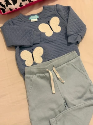 Blå quiltad med fjärilar från Pekkle🪻 - Supersöt blå quiltad väst från Pekkle med två stora broderade vita fjärilar framtill. Den  har rund hals, ribbade muddar och ett mjukt, vadderat material som känns riktigt mysigt. Perfekt för dig som vill ha något gulligt och bekvämt.