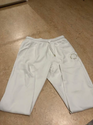 Vita mjukisbyxor från Hugo Boss - Säljer ett par vita mjukisbyxor från Hugo Boss med elastisk midja och snörning. Byxorna har fickor på sidorna och en diskret BOSS-logga tryckt på låret. Sköna och stilrena, perfekta för chill dagar eller streetwear-look.