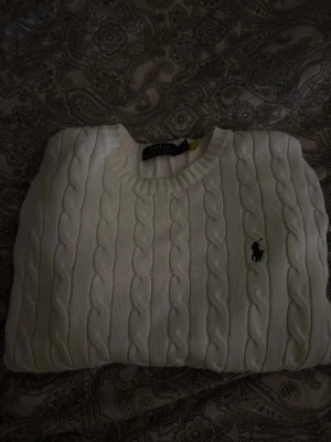 Vit kabelstickad tröja Polo Ralph Lauren - Kabelstickad vit tröja från Polo Ralph Lauren med klassisk rund halsringning och broderad logga på bröstet. Tröjan har långa ärmar och ribbade muddar vid ärmslut och nederkant. Perfekt för lager på lager under höst och vinter.