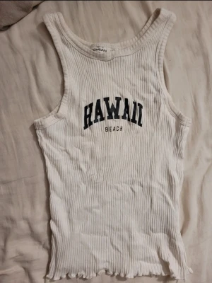 Vit ribbad linnetopp Hawaii Beach - Säljer en vit ribbad linnetopp från KappAhl med svart text 'HAWAII BEACH' framtill. Toppen är ärmlös och har en croppad passform med vågig kant nertill. Perfekt för sommardagar och strandhäng.