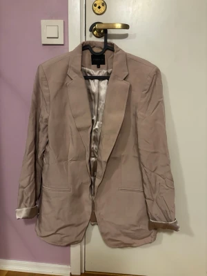 Beige kavaj från STOCKH LM - Säljer en stilren beige kavaj från STOCKH LM i storlek 42. Kavajen har klassisk krage, två fickor fram och är helfodrad med glansigt tyg. Perfekt för dig som vill ha en enkel men snygg look.