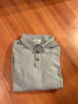 Ljusblå ullpiké från COS, slim fit - Säljer en stilren ljusblå/grå pikétröja från COS i slim fit. Tröjan är tillverkad i 100% ull, har lång ärm, klassisk krage och knäppning med tre knappar framtill. Perfekt för dig som gillar minimalistisk och clean stil. 