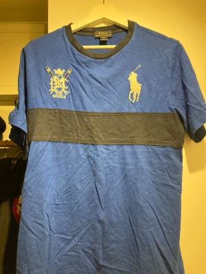 Blå Polo Ralph Lauren t-shirt XL - Blå t-shirt från Polo Ralph Lauren i storlek XL med svart band över bröstet, gul broderad polospelare och emblem framtill samt stor gul siffra 3 på ärmen. Klassisk rund halsringning med svart kant. Skön och mjuk bomullskvalitet.