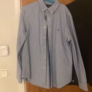 Ljusblå skjorta från Ralph Lauren - Klassisk ljusblå skjorta från Ralph Lauren med button-down krage och broderad logga på bröstet. Skjortan har långa ärmar och knäppning framtill. Perfekt för en clean och stilren look.