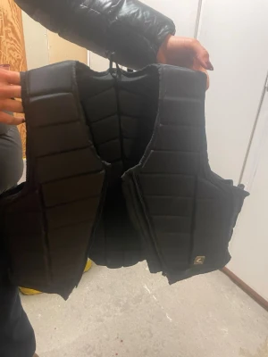 Svart quiltad säkerhetsväst XS - Säljer en svart quiltad säkerhetsväst från Z i storlek XS. Västen har en vadderad design med tydliga sömmar och breda axelband. Perfekt för ridning och ger bra skydd över rygg och bröst. Stängs med kardborre i sidan.