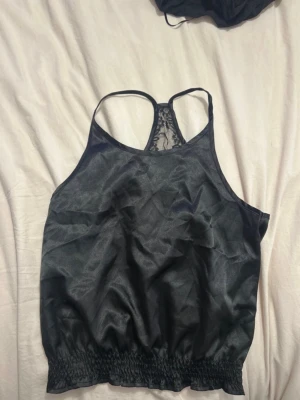 Svart linnetopp med spetsdetalj - Säljer en svart linnetopp med tunna axelband och snygg spetsdetalj i ryggen. Toppen har resår i nederkanten för en skön passform och är tillverkad i ett glansigt, tunt material som känns lätt mot huden.