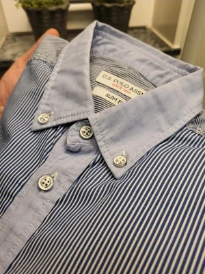 Randig blå skjorta från U.S. Polo Assn. S - Säljer en slim fit skjorta från U.S. Polo Assn. i blått och vitt med tunna ränder. Skjortan har klassisk krage, broderad logga på bröstet och vita knappar framtill. Tillverkad i bomull och har lång ärm. Perfekt för dig som gillar stilrena detaljer.