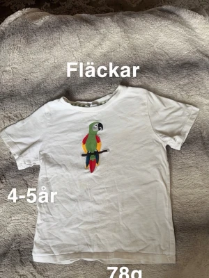 Vit t-shirt med papegojtryck - Vit t-shirt med färgglatt papegojtryck på bröstet. Klassisk rund hals och korta ärmar. Perfekt för dig som gillar djurmotiv och vill ha en enkel men lekfull stil.🌸 Fläckar. Storlek 4-5år =110