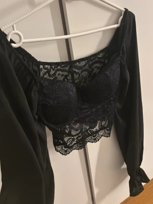 Svart spetsig korsettopp - Supersnygg svart korsettopp från SHEIN i storlek XS med långa puffärmar och detaljer i spets. Toppen har croppad passform, v-ringning och transparent spets över bysten och magen. Gåva vid köp!🎁