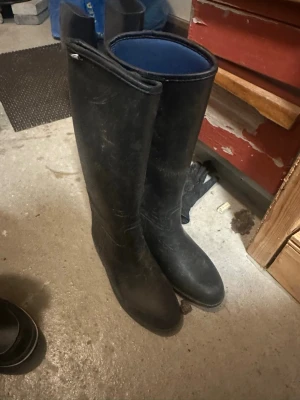 Svarta höga stövlar från Tretorn - Säljer ett par klassiska svarta höga stövlar från Tretorn. De är tillverkade i slitstark gummi med blått foder på insidan och har en rund tå samt platt sula. Perfekta för regniga dagar eller utomhusaktiviteter.