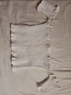 Vit smockad bandeau topp - Säljer en vit bandeau topp med smockad struktur och volangkant upptill och nedtill. Toppen är axelbandslös och har en stretchig passform som sitter tight mot kroppen. Perfekt för sommardagar och enkel att matcha med olika stilar.