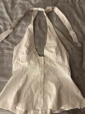 Vit randig halterneck topp från Zara - Supersnygg halterneck topp från Zara i vitt med tunna beige ränder upptill. Toppen har knyt i nacken, knappar framtill och lätt utsvängd nederdel. Perfekt till sommarens alla outfits och ger en chic vibe med sin omlottkänsla och djupa urringning. Liten i storleken!!