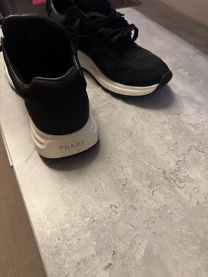 Svarta sneakers från Prada - Säljer ett par svarta sneakers från Prada med vit sula och klassisk logga på både plös och häl. Skorna har snörning och en stilren design med diskreta detaljer i svart och vitt. Materialet är en mix av skinn och syntet som ger en lyxig känsla.