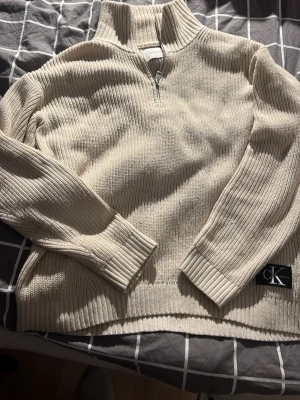 Beige stickad half zip från Calvin Klein - Säljer en beige stickad tröja från Calvin Klein Jeans med half zip och hög krage. Tröjan har ribbade muddar, lång ärm och en snygg CK-logga nertill. Perfekt för dig som gillar stilrena och bekväma plagg. Storlek 14 barn men väldigt stor i storleken. Skulle passa som en ”vanlig” S
