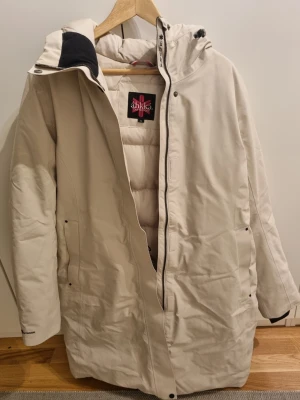 Beige vinterparkas från Áhkká - Säljer en beige vinterparkas från Áhkká med huva och dragkedja. Jackan är tillverkad i vind- och vattentätt material med tejpade sömmar och andasfunktion. Perfekt för kalla dagar och har en clean, stilren look med praktiska fickor.