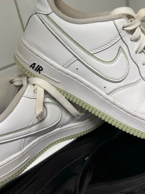 Nike Air Force 1 vita sneakers - Klassiska Nike Air Force 1 sneakers i vitt skinn med ljusgrön detalj på sulan och swoosh. Skorna har rund tå, perforeringar på tån för ventilation och snörning framtill. Tidlös modell som passar till det mesta och är superlätta att styla.