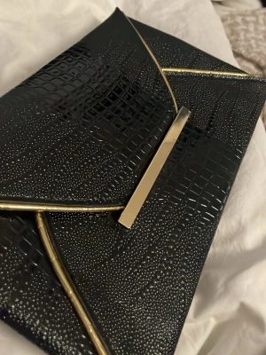Svart kuvertväska med guldiga detaljer - Snygg svart kuvertväska i glansigt material med krokopräglat mönster och guldiga metallkanter. Väskan har en stilren, modern look och stängs med ett metallspänne framtill. Perfekt accessoar för att lyfta din outfit.