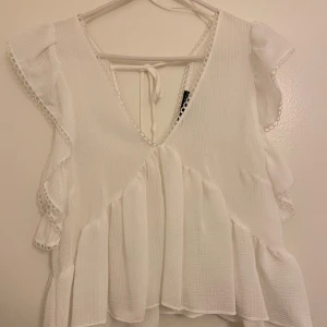 Vit blus från Zara - Säljer en söt vit blus från Zara med volanger och knytning i ryggen. Blusen har en v-ringad design och är ärmlös med fina spetsdetaljer längs kanterna. Perfekt för en somrig look!