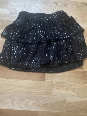 Svart glittrig volangkjol - Säljer en svart kortkjol med glittriga paljetter och dubbla volanger. Kjolen har en riktigt festlig vibe och är perfekt för dig som vill sticka ut. Elastisk midja för skön passform och skimrande material som fångar ljuset.