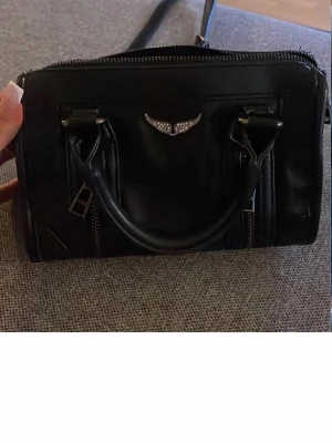 Zadig väska liknande - Cool svart handväska i skinnimitation med två handtag och avtagbar axelrem. Väskan har silvriga dragkedjor framtill och en snygg vingdetalj i metall med små stenar. Perfekt storlek för mobilen, plånboken och lite smink.