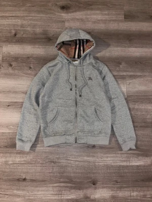 Burberry grey zip hoodie, check lining, designer hoodie - Grey Burberry zip hoodie i mjukt och skönt material med klassiskt Burberry-rutigt foder i huvan. Tidlös och lättmatchad. I fint skick med normalt bruksslitage, se bilder. Kommer från ett djur- och rökfritt hem. Skickas samma dag eller senast dagen efter köp. Fråga gärna om fler bilder eller info!