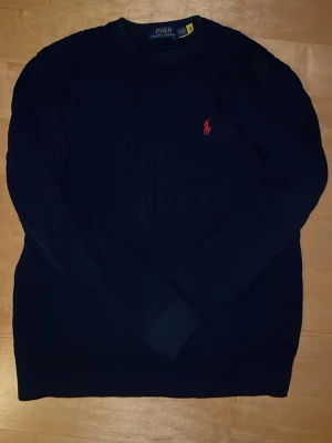 Mörkblå stickad tröja Polo Ralph Lauren - Mörkblå stickad tröja från Polo Ralph Lauren med klassisk röd logga broderad på bröstet. Rund halsringning och långa ärmar. Perfekt till höst och vinter för en clean och tidlös look.