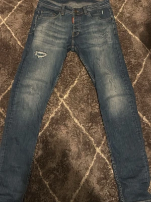 Blå slim fit jeans från Dsquared2 - Säljer ett par blå slim fit jeans från Dsquared2 med slitningar och distressed detaljer på framsidan. Klassisk femficksmodell med låg midja och snygg tvätt. Jeansen har en röd Dsquared2-label bak i linningen och är tillverkade i stretchig denim för skön passform. Längden på jeansen är 33 och midjan 31