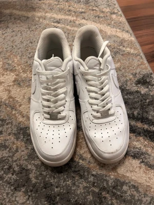 Nike Air Force 1 vita sneakers - Klassiska Nike Air Force 1 sneakers i helvitt skinn med rund tå och snörning. Snygg och tidlös modell med perforerade detaljer på tån och Nike-logga på sidan och hälen. Passar perfekt till streetwear och casual outfits.