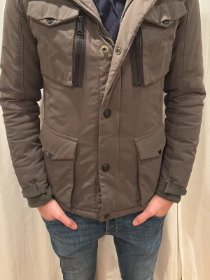 Grön pufferjacka med fickor och dragkedjor - Säljer en grön pufferjacka med fyra stora fickor framtill, två med lock och två med dragkedja. Jackan har knappstängning och hög krage. Perfekt för kyliga dagar och har en clean, modern look. Från märket Schott N.Y.C och nyspris är ungefär 2200