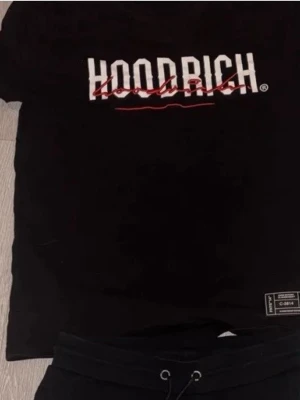 Svart HOODRICH t-shirt med tryck - Svart t-shirt från HOODRICH med stort vitt och rött tryck på bröstet. Klassisk passform och rund hals. T-shirten har en liten etikett nertill och är perfekt för en streetwear-look.