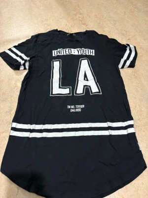 Svart t-shirt Jack & Jones LA-print - Svart t-shirt från Jack & Jones CORE med stort vitt LA-tryck och texten UNITED:YOUTH på bröstet. T-shirten har vita ränder på ärmarna och nedtill, samt rundad nederkant. 