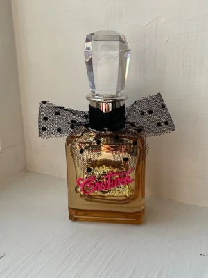 Juicy Couture parfym med rosett - Snygg parfym från Juicy Couture med dekorativ svart rosett och guldiga detaljer. Flaskan har en lyxig känsla och passar perfekt för dig som vill sticka ut. Ungdomlig söt doft, 50ml. Flaskan är full och endast testad 🩷