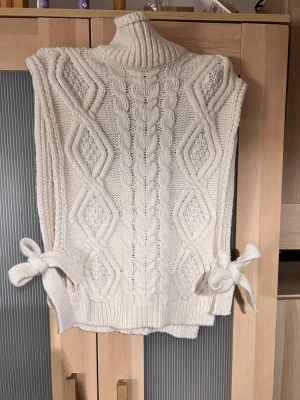 Vit stickad väst med knyt i sidan - Säljer en chunky stickad väst i offwhite med polokrage och snygga kabelstickade detaljer framtill. Västen har breda axlar och knytband i sidorna för en cool touch. Perfekt att slänga över en skjorta eller långärmad tröja för extra lager. Står inget märke eller storlek på den men den passar dem som söker kläder i stl XS/S. Den har lite lösa tråd som man ser på bilden men inget som syns mycket m. Den sitter otroligt fint på. 