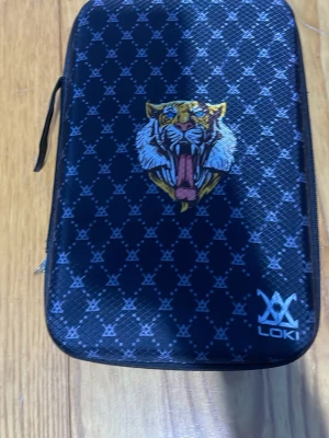 Pingisväska med tigertryck från LOKI - Snygg svart pingisväska från LOKI med blått mönster och ett stort tigertryck på framsidan. Väskan har dragkedja och plats för racket och boll. Perfekt för att ta med till träningen eller matchen.