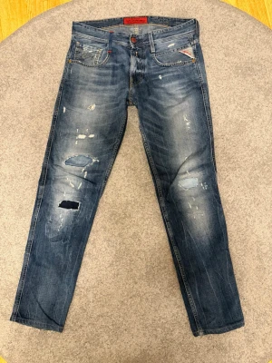 Replay jeans med slitningar - Säljer ett par blå unika Replay jeans med slitningar och patchade detaljer på benen. Jeansen har klassisk femficksmodell och snyggt tvättad denim. Märkesdetaljer på fickorna och bak med Replay-lapp i läder. Storlek: W29, L32. Vid funderingar eller liknande är det bara att höra av sig! 
