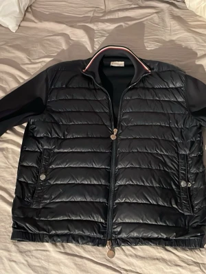 Svart pufferjacka från Moncler - Roligt fet Svart pufferjacka från Moncler med quiltad framsida och släta ärmar. Jackan har dragkedja framtill, ståkrage med rödvit rand och två fickor med tryckknappar. Klassisk Moncler-logga på ärmen och sportig look. Helt äkta. Nypris 12050 kr. Mitt pris 5000. Säljer den eftersom den inte är till användning längre.