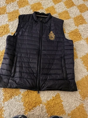 Mörkblå väst  från Morris Gent's - Säljer en mlrkblå quiltad väst från  Morris Gent's med guldfärgat broderat emblem på bröstet. Västen har hög krage, dragkedja framtill och två sidofickor. 