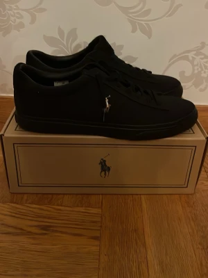 Svarta Polo Ralph Lauren sneakers - Snygga svarta sneakers från Polo Ralph Lauren, modell Sayer-NE. Skorna har en stilren design med Polo-logga på sidan och brun gummisula med Polo-tryck undertill. Ovandelen är tillverkad av textil och läder, med syntetfoder och snörning framtill. Helt oanvända, säljer dom pga att dom är för stora