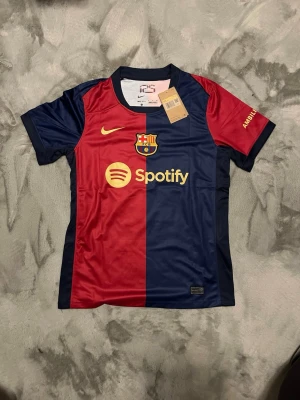 Barca matchtröja Lamine Yamal 19 - Säljer en officiell FC Barcelona matchtröja från Nike med Lamine Yamal och nummer 19 på ryggen. Tröjan har klassiska röda och marinblå fält, gul Nike-logga och Spotify som sponsor framtill. Tillverkad i lätt polyester, perfekt för fotbollsplanen.