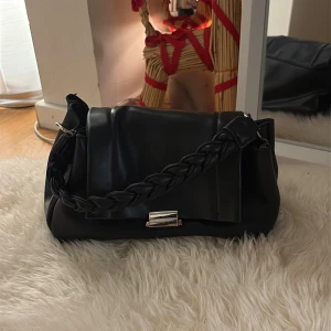 Svart axelväska med flätat handtag - Snygg svart axelväska i skinnimitation med ett flätat handtag och silverfärgat spänne framtill. Väskan har en mjuk, rundad form och stilren design som passar till många outfits.