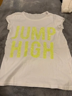 Vit t-shirt med paljetter och text - Vit t-shirt i bomull med korta ärmar och rund hals. Framsidan har stora neongula bokstäver i paljetter som stavar 'JUMP HIGH'. Perfekt för dig som gillar statement-plagg och vill sticka ut lite extra.