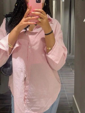 Ljusrosa randig skjorta - Säljer en ljusrosa skjorta med tunna vita ränder. Skjortan har klassisk krage, knäppning framtill och långa ärmar med breda manschetter. Passformen är loose och materialet känns som bomull, perfekt för en chill och avslappnad stil.